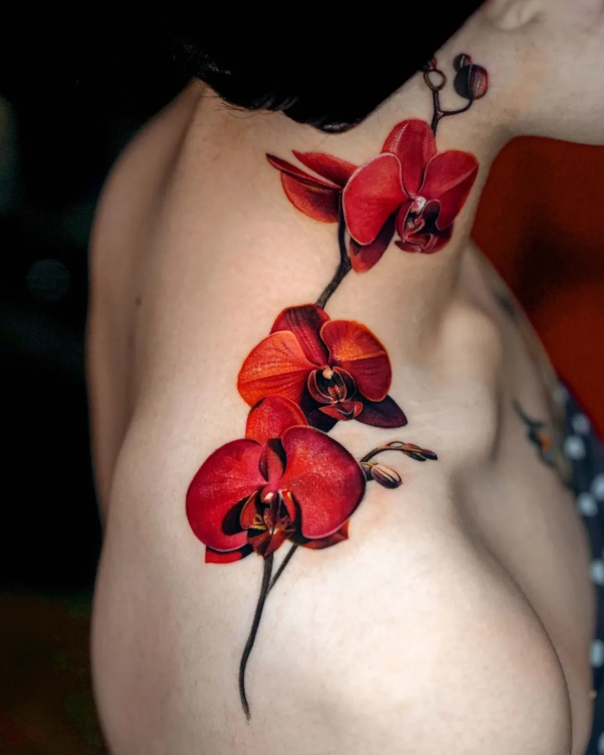 A close-up of a shoulder and upper chest tattoo showing three open red Phalaenopsis orchid blooms and two buds on a dark stem that curves from the base of the neck across the clavicle, rendered in shades of deep red and burgundy.