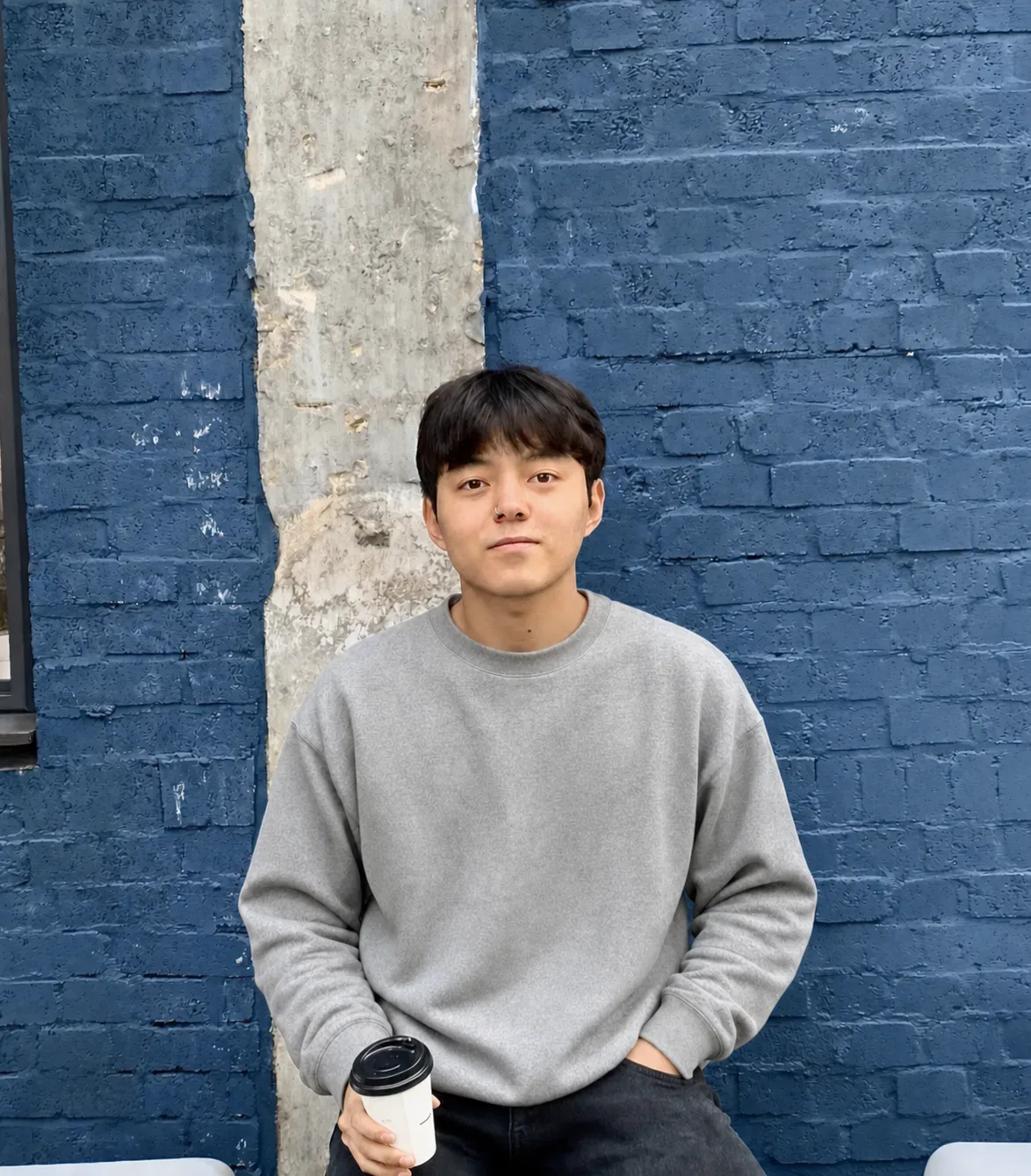A young East Asian man with dark hair wearing a light gray crewneck sweatshirt and dark jeans stands in front of a blue brick wall with a vertical concrete stripe, looking directly at the camera and holding a white disposable coffee cup.