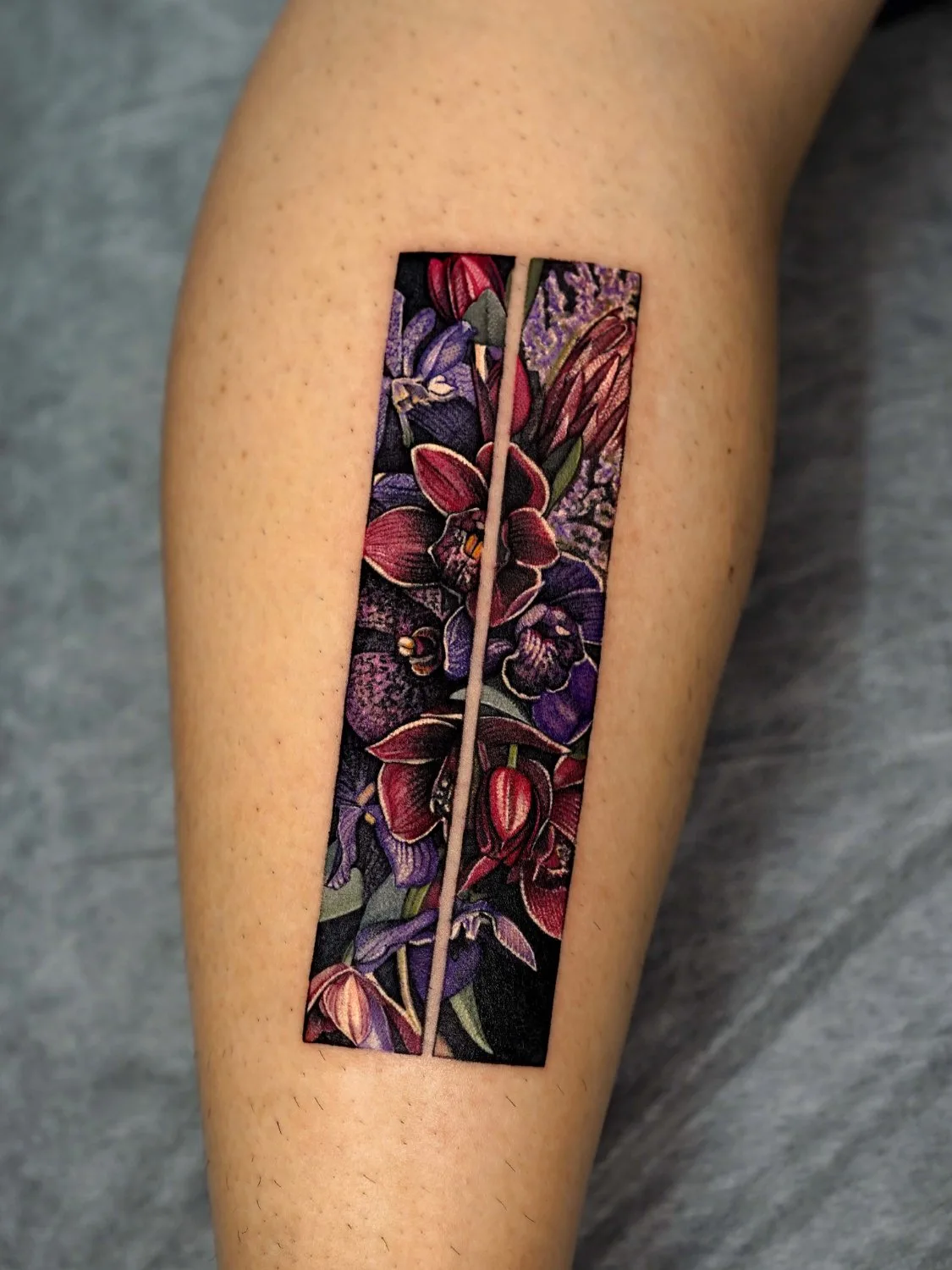 A close-up of a leg tattoo consisting of two narrow vertical rectangular panels side by side, filled with richly colored deep-red and purple orchid-like flowers, pointed buds, and green leaves on a very dark background.