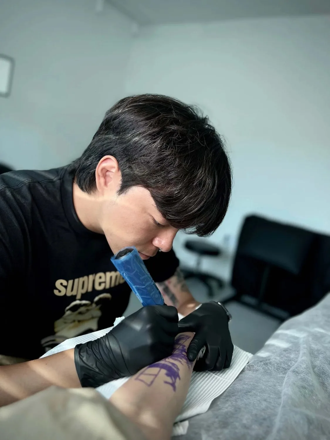 A young man with dark styled hair wearing a black Supreme t-shirt and black nitrile gloves leans over a client's forearm, which rests on a white paper towel and is covered in a purple geometric stencil, as he applies a tattoo.