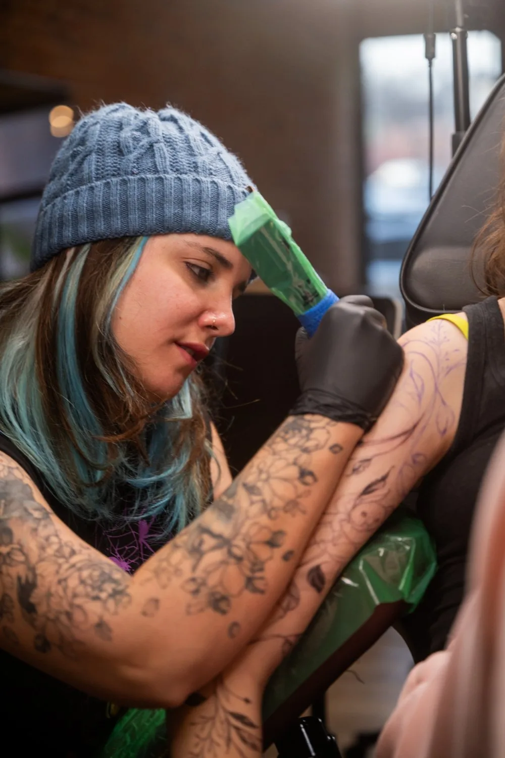 A tattoo artist with blue-streaked hair and a grey beanie leans over a client's arm, tattooing a light purple floral stencil with a green-wrapped tattoo machine, her own floral black-ink tattoos visible on her arms.