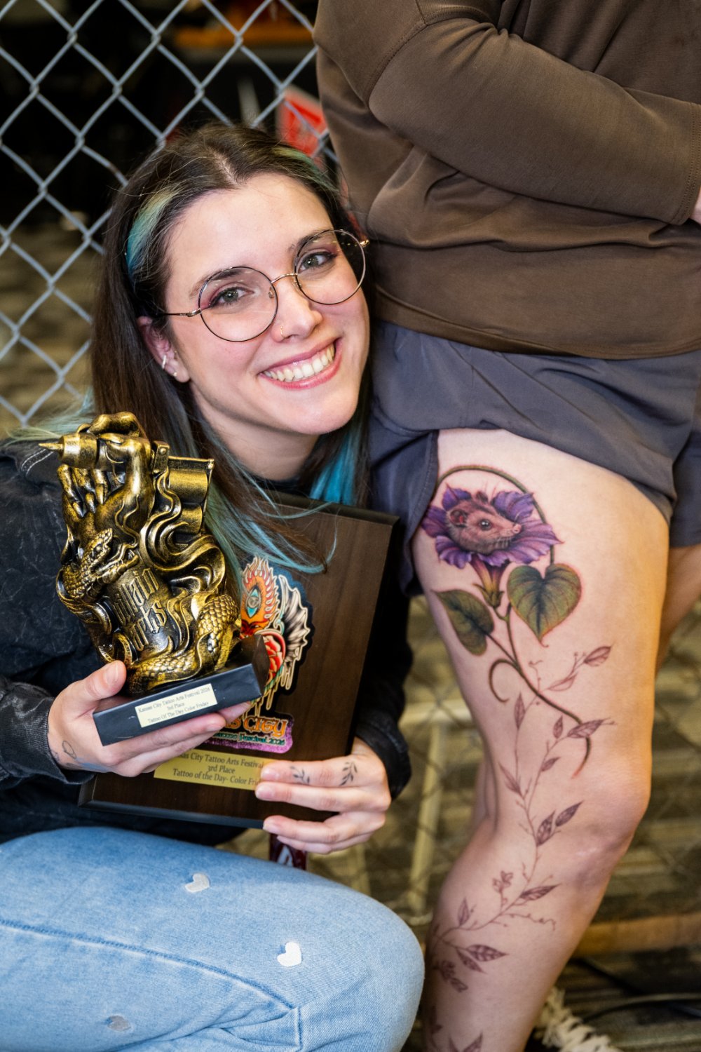 A smiling woman with blue-streaked hair in a ponytail and round glasses holds a golden claw-shaped trophy and a wooden award plaque; to her right, a leg displays a detailed tattoo of a purple flower with a small animal face at its center.