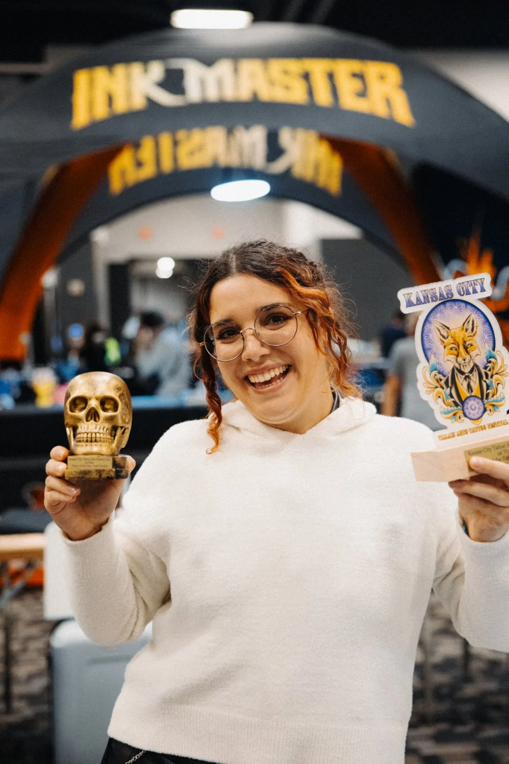 A woman with curly reddish-brown hair and round glasses laughs while holding a small golden skull trophy in one hand and a wooden award plaque featuring a fox-in-suit emblem and Kansas City branding in the other, inside a convention hall.