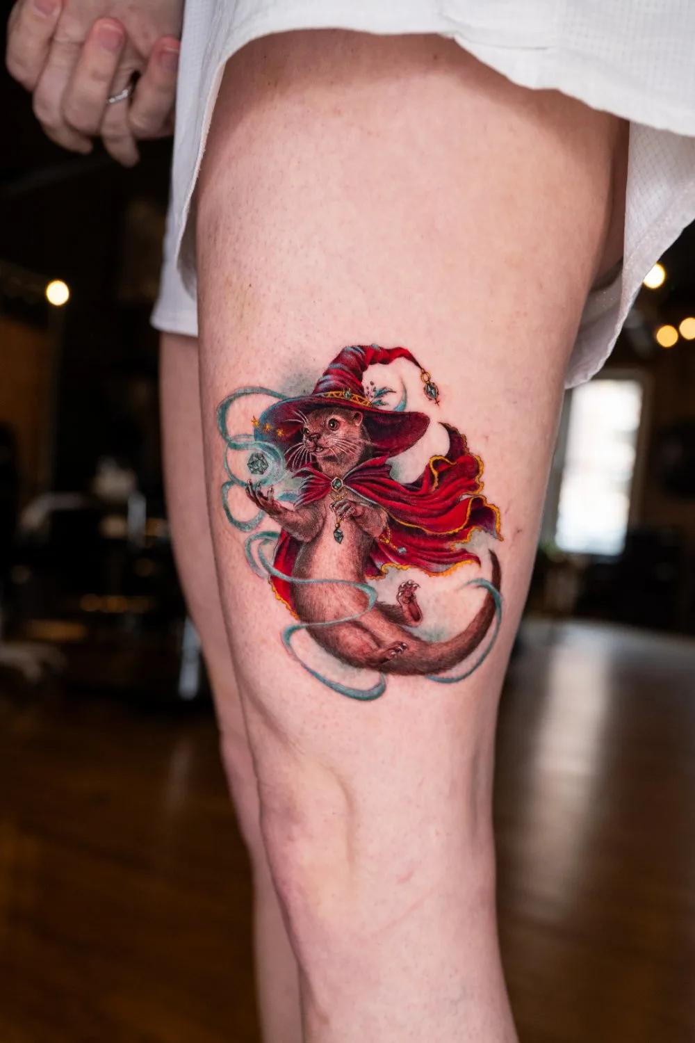 Close-up of a thigh tattoo showing a detailed otter dressed as a wizard, wearing a red pointed hat with stars and a flowing red cape, one paw raised and the other holding a glowing orb surrounded by teal magical energy.