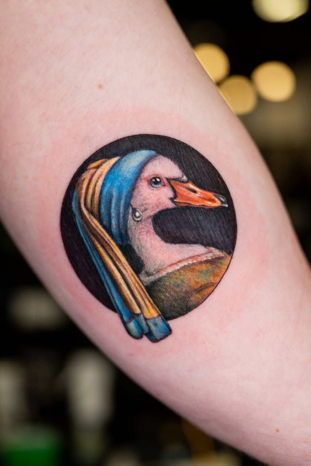 Circular tattoo depicting a goose in profile wearing a blue and gold headscarf and a pearl earring, rendered in rich color realism against a dark background in direct homage to Vermeer's Girl with a Pearl Earring.