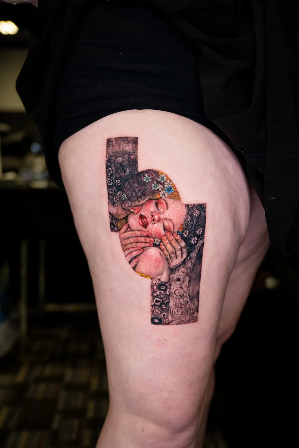 Close-up of a thigh tattoo depicting two figures kissing in the style of Klimt's The Kiss, with intricate decorative patterning, soft skin tones, and red lips, framed within a slightly curved rectangular border.