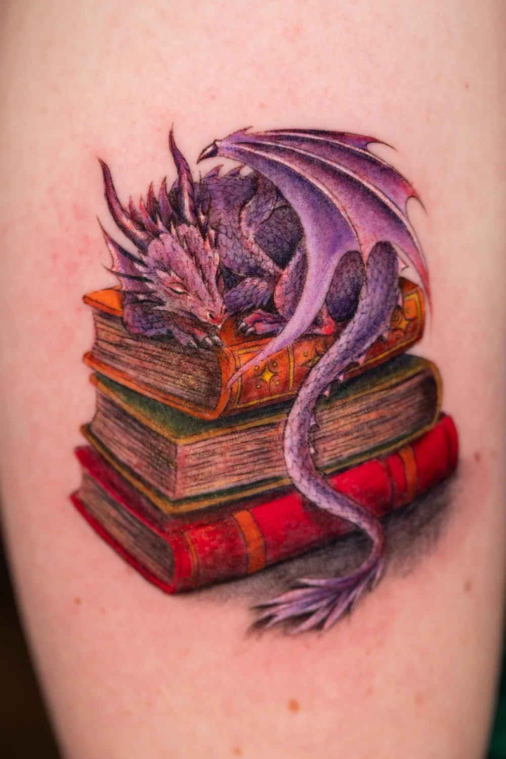 Close-up of a tattoo showing a small dragon with purple and lavender scales and translucent wings sleeping on top of three stacked books with red, green, and gold bindings, its tail draped around the stack.