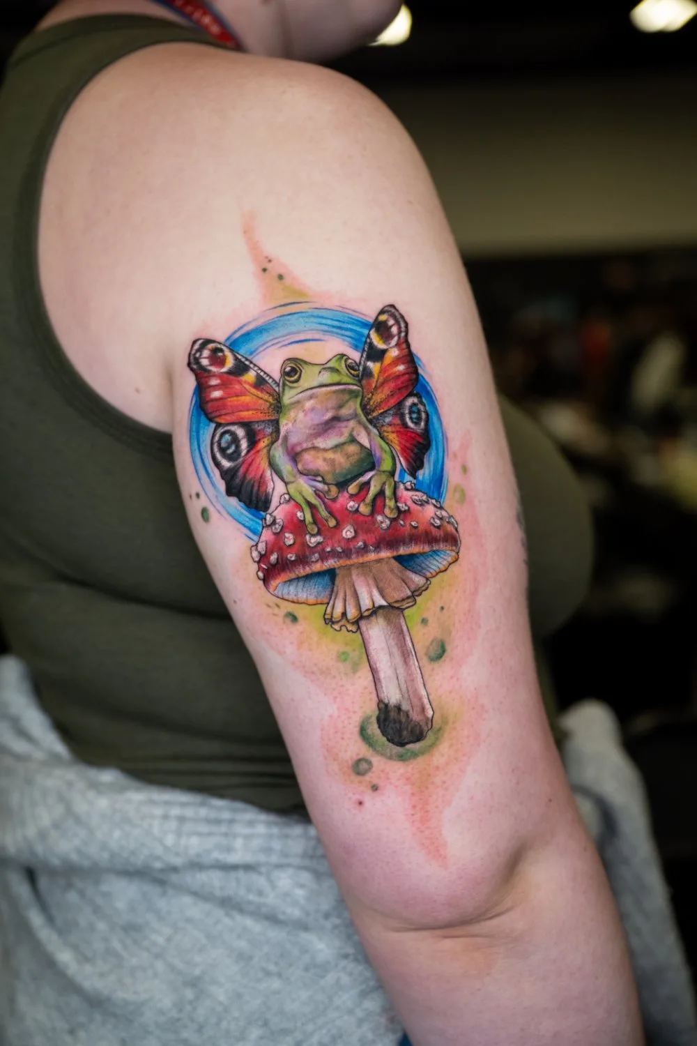 Upper arm tattoo of a green frog with large butterfly wings in red, orange, and black resting on a red and white spotted mushroom, encircled by a bright blue disc with soft yellow and orange washes around it.
