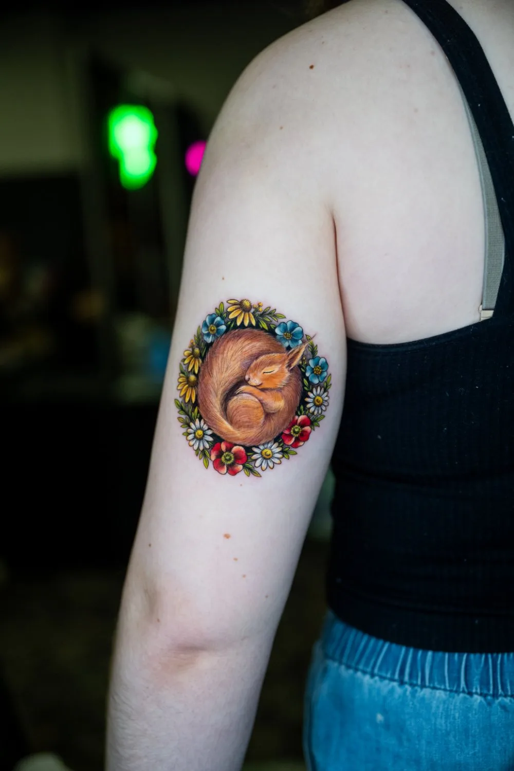 Upper arm tattoo of a red squirrel sleeping with its tail wrapped around its body, surrounded by a detailed circular wreath of white daisies, small blue flowers, red anemones, and green leaves.
