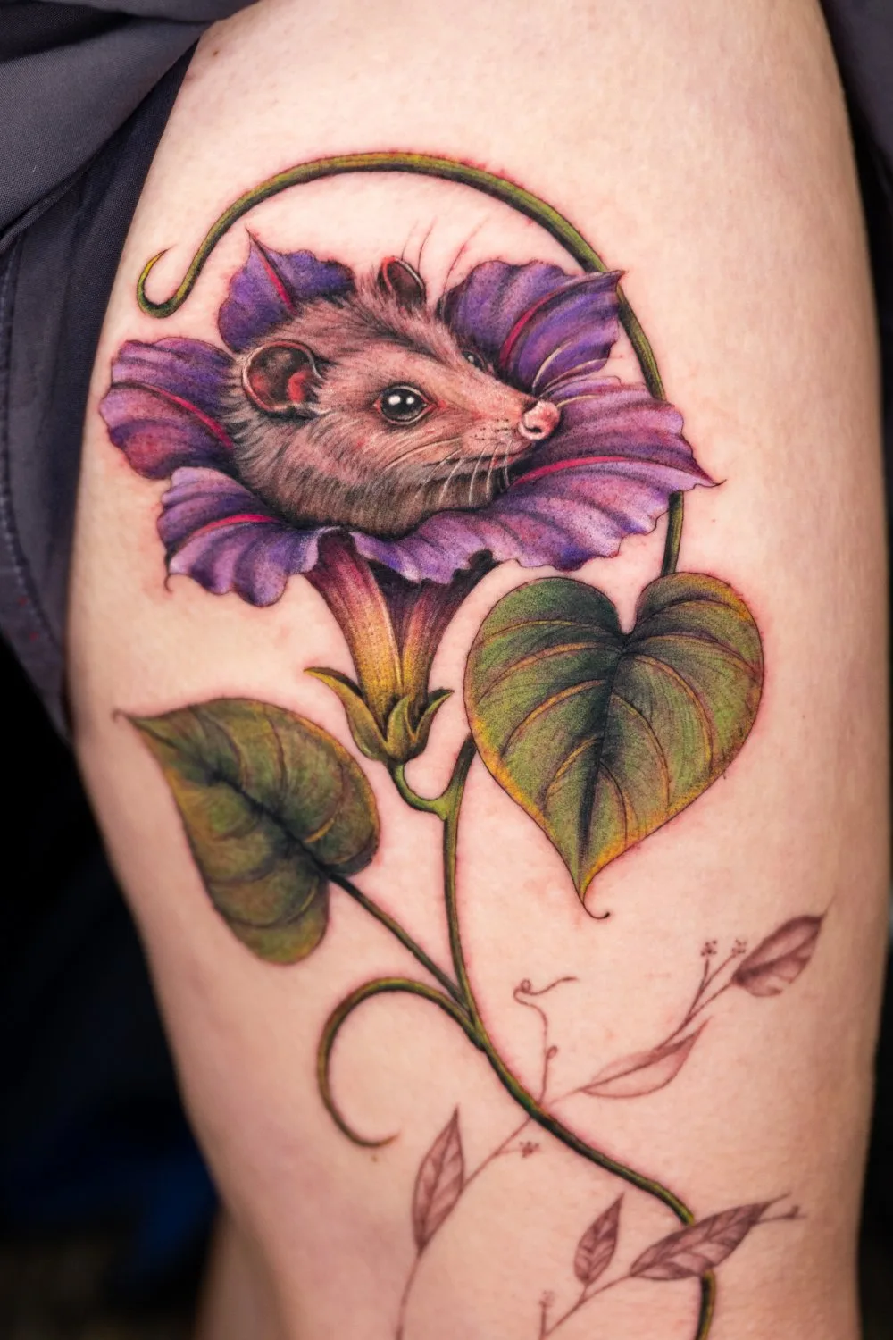 Thigh tattoo showing a small light-brown mouse peeking out from the center of a large purple flower with dark veined petals, attached to a thick green vine with two large heart-shaped leaves and smaller faded seed pods below.