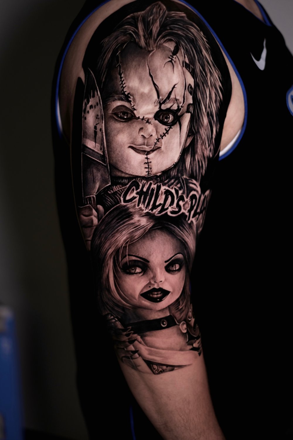 A black and grey photorealistic tattoo on an upper arm showing Chucky's stitched, cracked face holding a knife at the top and Tiffany Valentine's face with blonde hair and studded collar below, with the text 'CHILD'S PLAY' in a distressed font between the two characters.