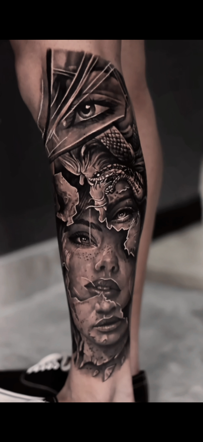 A black and grey hyperrealistic tattoo on a lower leg showing a woman's face fragmented as if breaking apart behind torn geometric shapes, with a detailed coiled snake positioned at the top of the design near the face, and a prominent eye visible through the angular fragments.