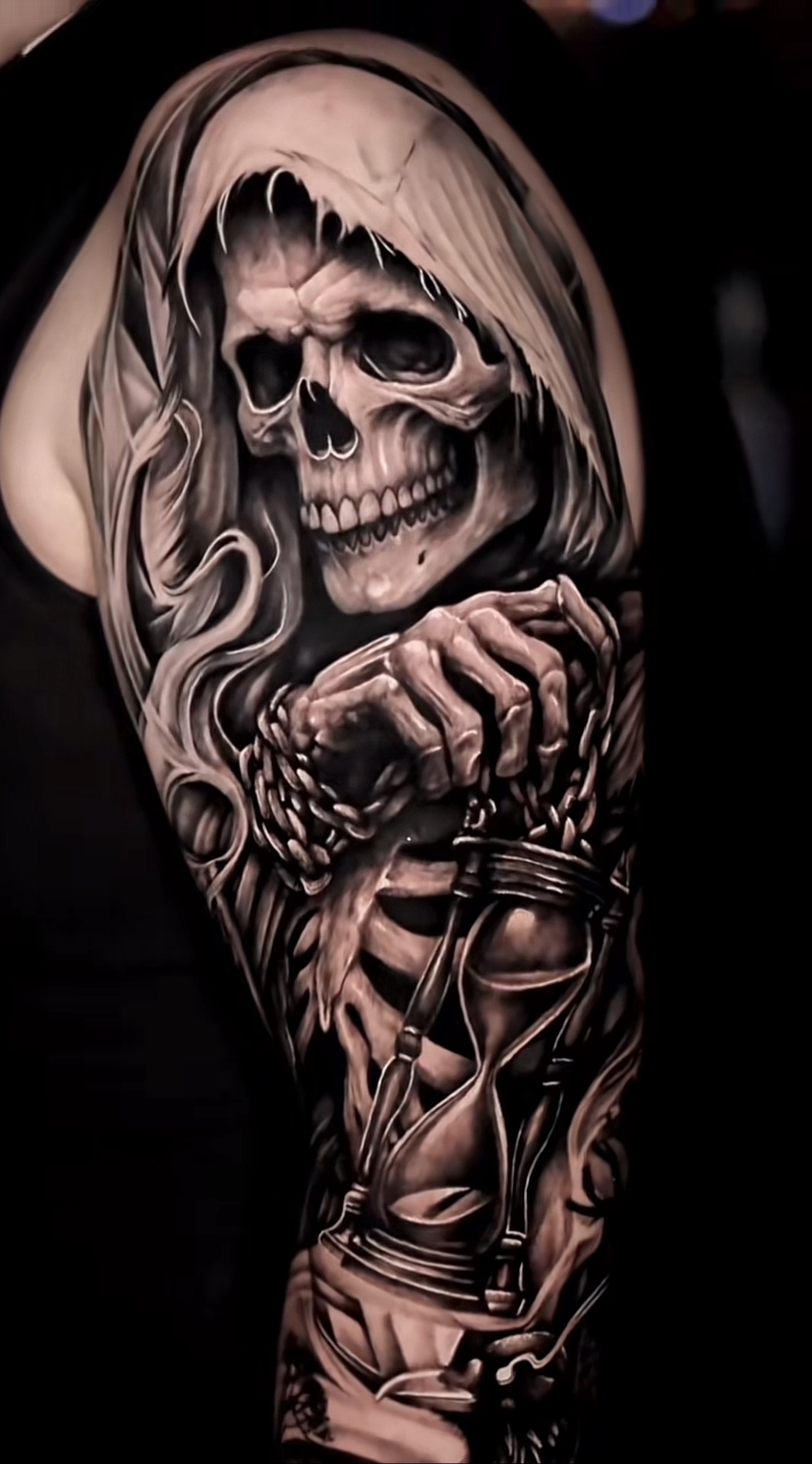 A black and grey photorealistic tattoo on an upper arm and shoulder depicting the Grim Reaper as a skull in a dark hood, with skeletal hands below gripping a chain attached to an hourglass with sand flowing through it. The rendering uses dramatic light and shadow to create a three-dimensional effect