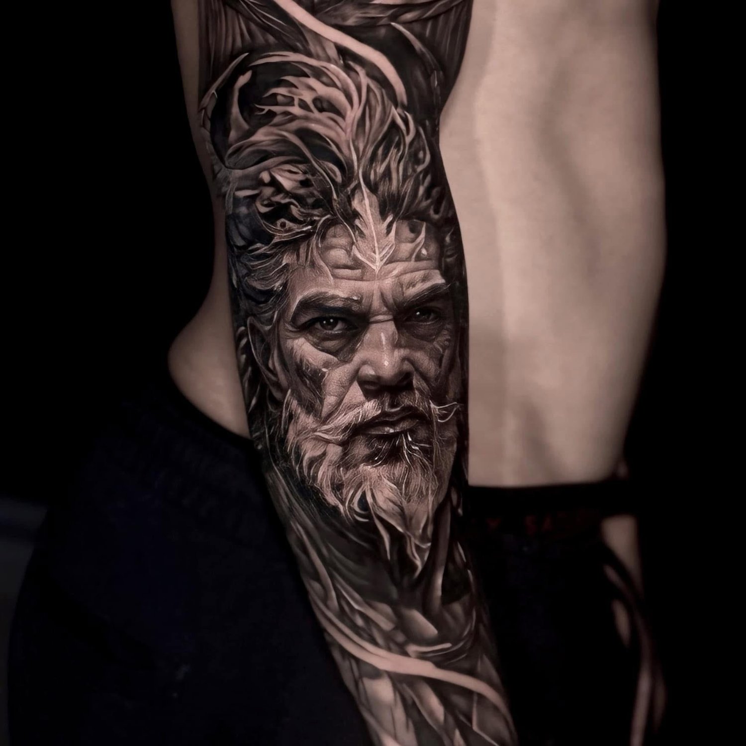 A black and grey hyperrealistic tattoo on an upper arm depicting the face of an elderly man with a full bushy beard, prominent wrinkles, deep-set eyes, and wild windswept hair. Abstract swirling lines extend around and behind the head against a dark background.