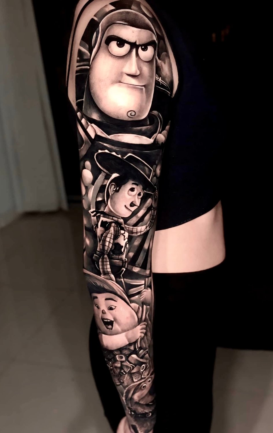 A black and grey full-sleeve tattoo on an arm showing Buzz Lightyear's helmeted face and determined expression at the top, Woody in cowboy attire holding his hat below, and Russell from Up in his scout uniform further down, all rendered in a highly realistic style with smooth shading and strong cont