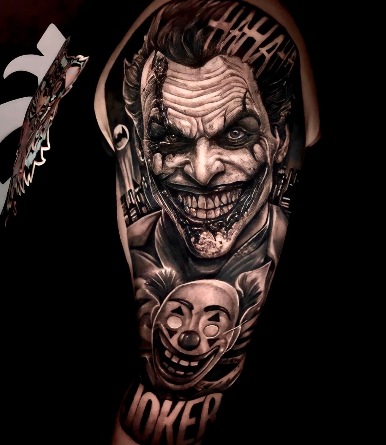 A black and grey hyperrealistic tattoo on an upper arm showing the Joker's sinister face with exaggerated grin and gaunt features in the upper portion, a smaller clown mask below, subtle cityscape elements in the background, and the word 'JOKER' in large lettering at the bottom.