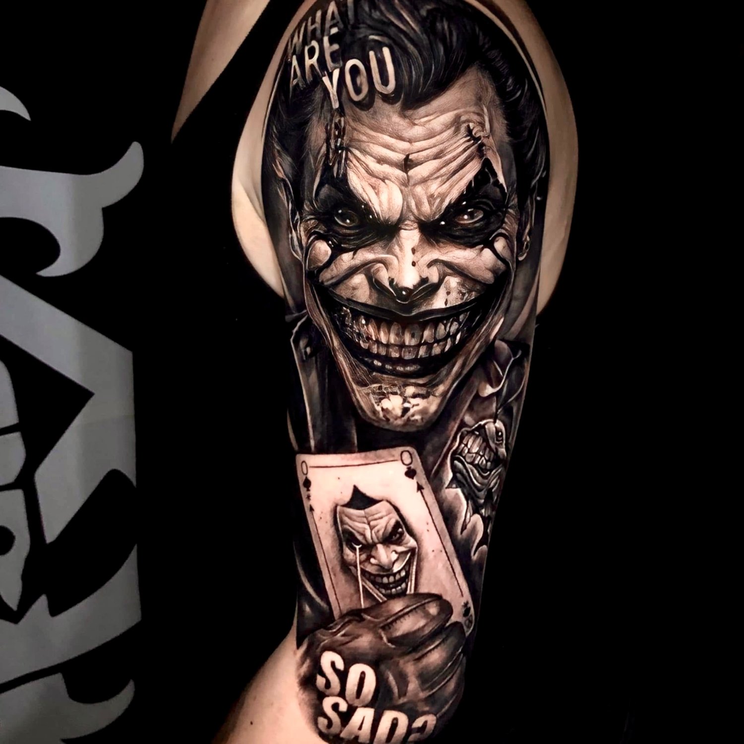 A close-up of a black and grey hyperrealistic Joker tattoo on an upper arm. The Joker's face fills the frame with a wide grin and dark eyes. He holds an Ace of Spades playing card featuring a small Joker face. Text reading 'WHAT ARE YOU' appears on his forehead and 'SO SAD' appears below on a dice-l