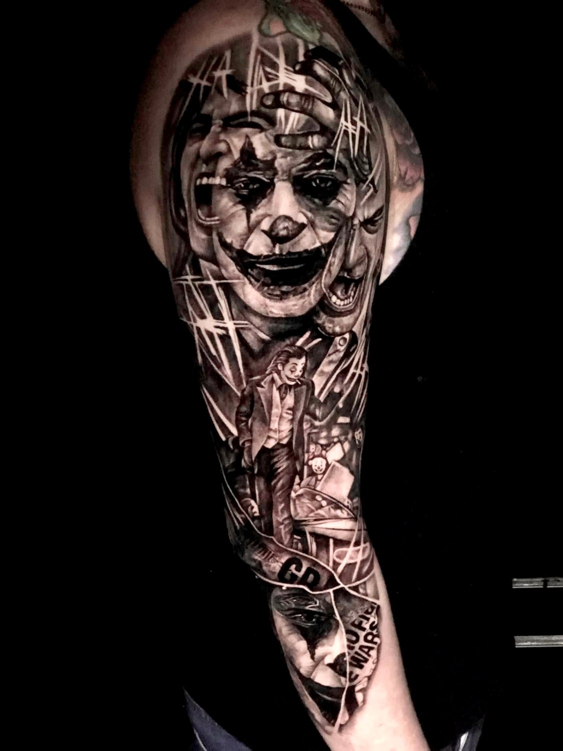 A black and grey realism full-sleeve tattoo featuring multiple overlapping and fragmented depictions of the Joker's face in various expressions across the upper arm and shoulder, a full-figure Joker in a suit holding a cigarette in the lower section, and scattered elements including playing cards, a