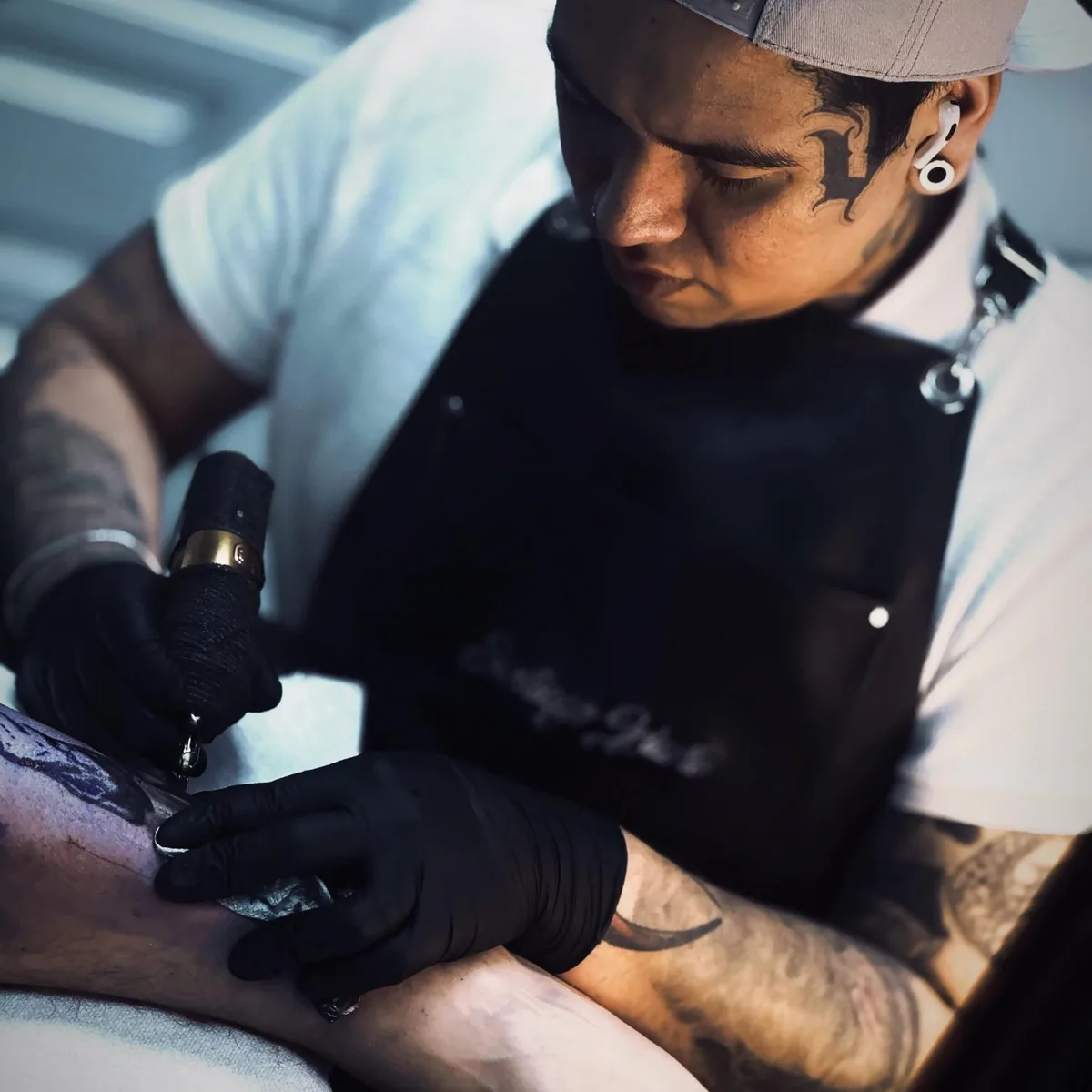 A tattoo artist wearing a grey baseball cap, black apron over a white t-shirt, and black nitrile gloves leans over a client's leg, holding a tattoo machine. The artist has facial tattoos and piercings. The depth of field is shallow, keeping the machine and leg in sharp focus.