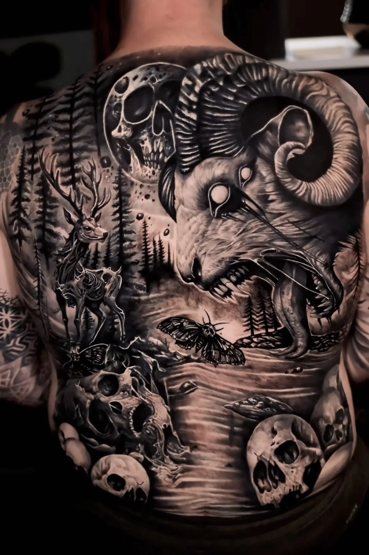 A full-back black and grey realism tattoo featuring a large demonic ram's head with spiraling horns at center, a skeletal deer to the left, a crescent moon with a skull impression above, multiple human skulls throughout, large moths, and a dense shadowy forest background.