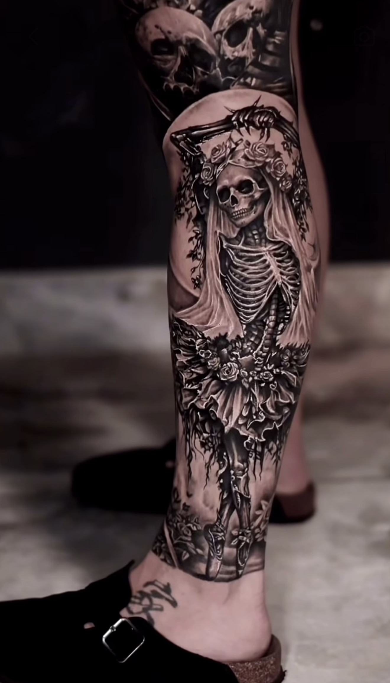 A black and grey realism tattoo running the full length of a leg, depicting a skeleton dressed as a bride with a delicate veil, floral wreath on the skull, and a layered tutu skirt adorned with flowers. The skeleton's legs are posed in ballet positions wearing pointe shoes. Additional skulls and dar