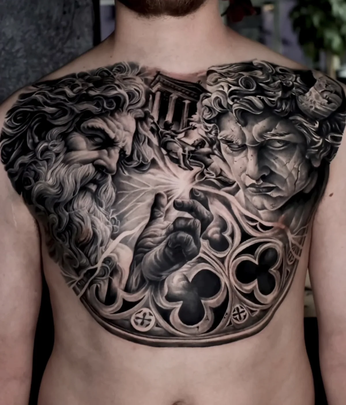 A black and grey realism chest tattoo showing a bearded god-like figure on the left and a cracked, horned figure on the right, a hand reaching toward a glowing light source between them, classical columns in the background, and an ornate gothic border along the bottom edge.