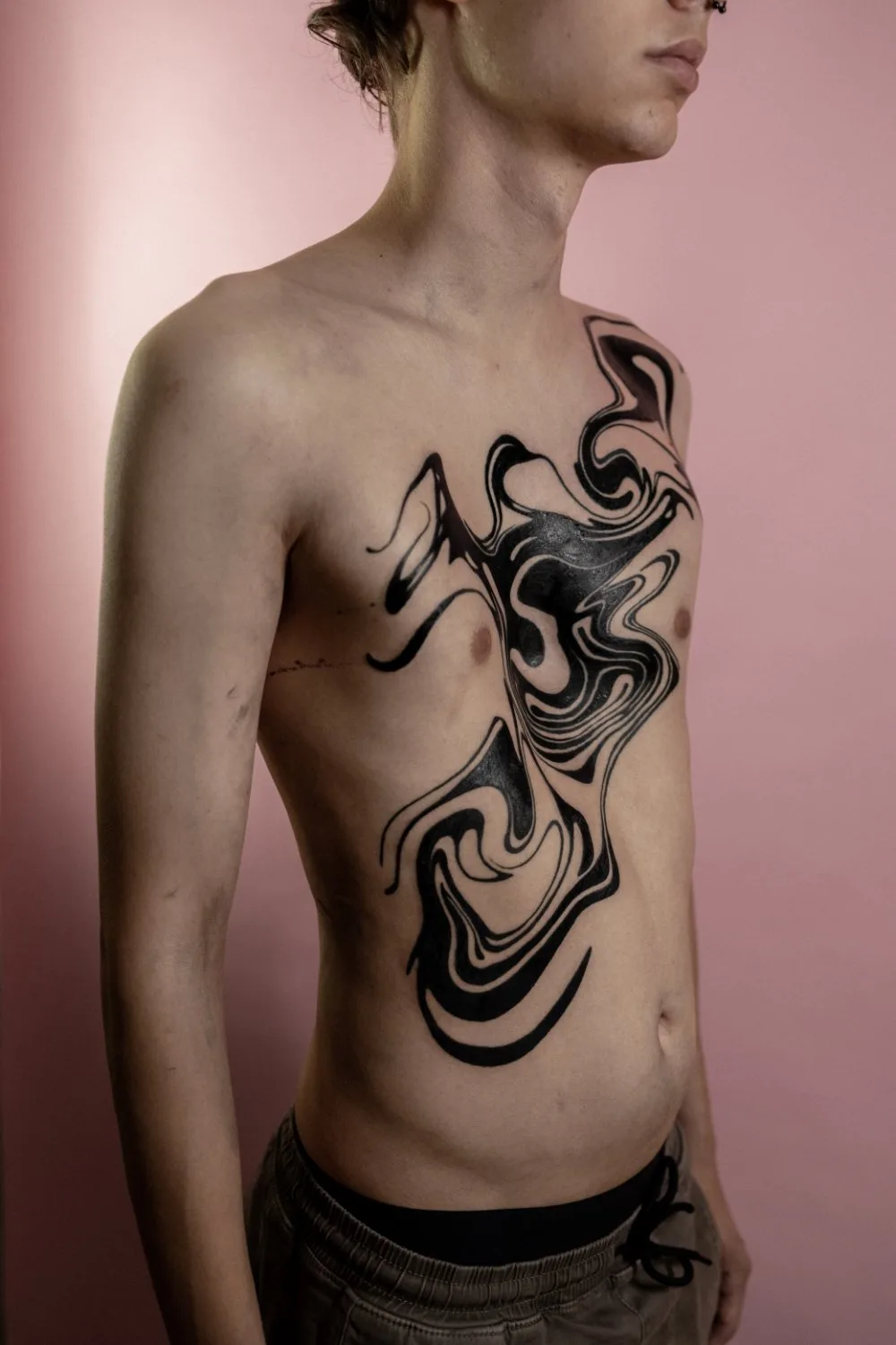 Close-up of a person's torso showing a large abstract black ink tattoo covering the chest and upper abdomen. The design features fluid, swirling lines of varying thickness against pale skin, with a soft pink background.