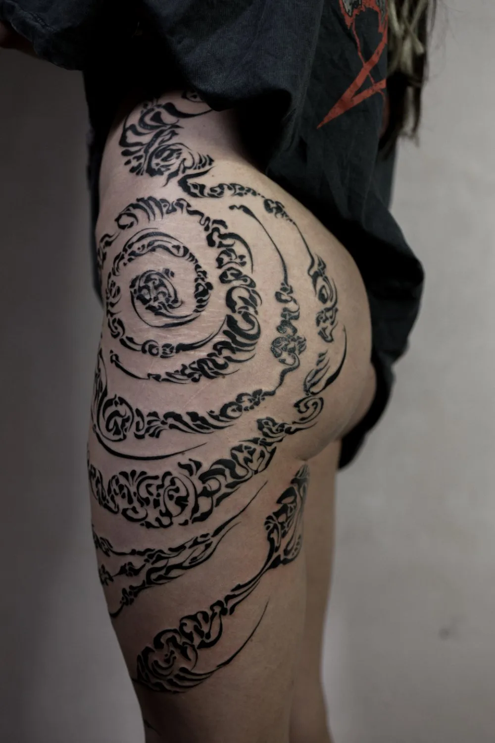 A person's thigh and hip area showing an intricate black ink tattoo. The design features swirling ornamental patterns with varying line weights, denser at the top and becoming more open and linear toward the lower portion of the thigh.