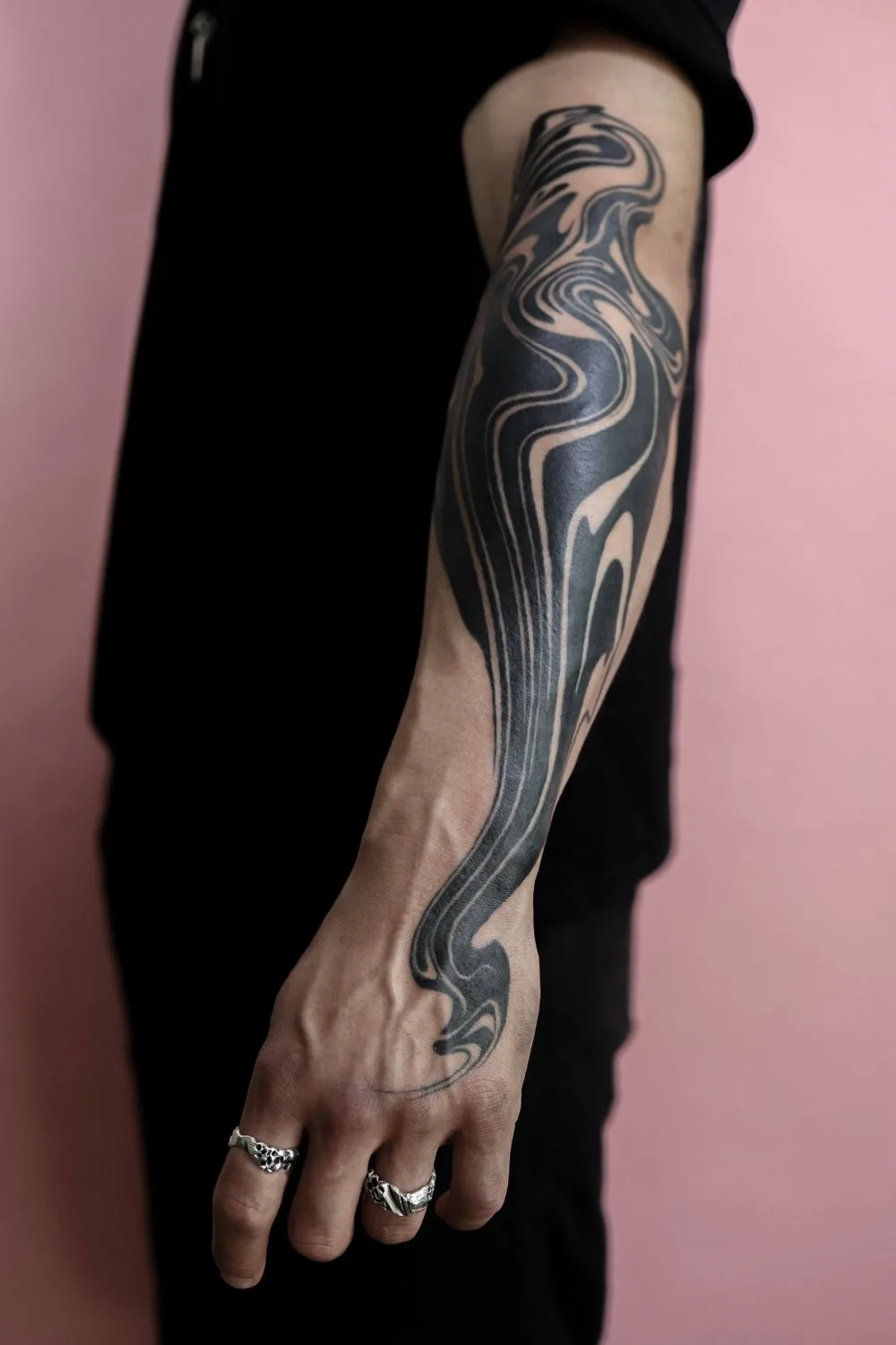 A close-up of a person's forearm and hand showing a bold abstract black ink tattoo. The design features swirling, fluid shapes with sharp edges and areas of negative space. Two silver rings are visible on the hand. The background is split between black fabric and soft pink.