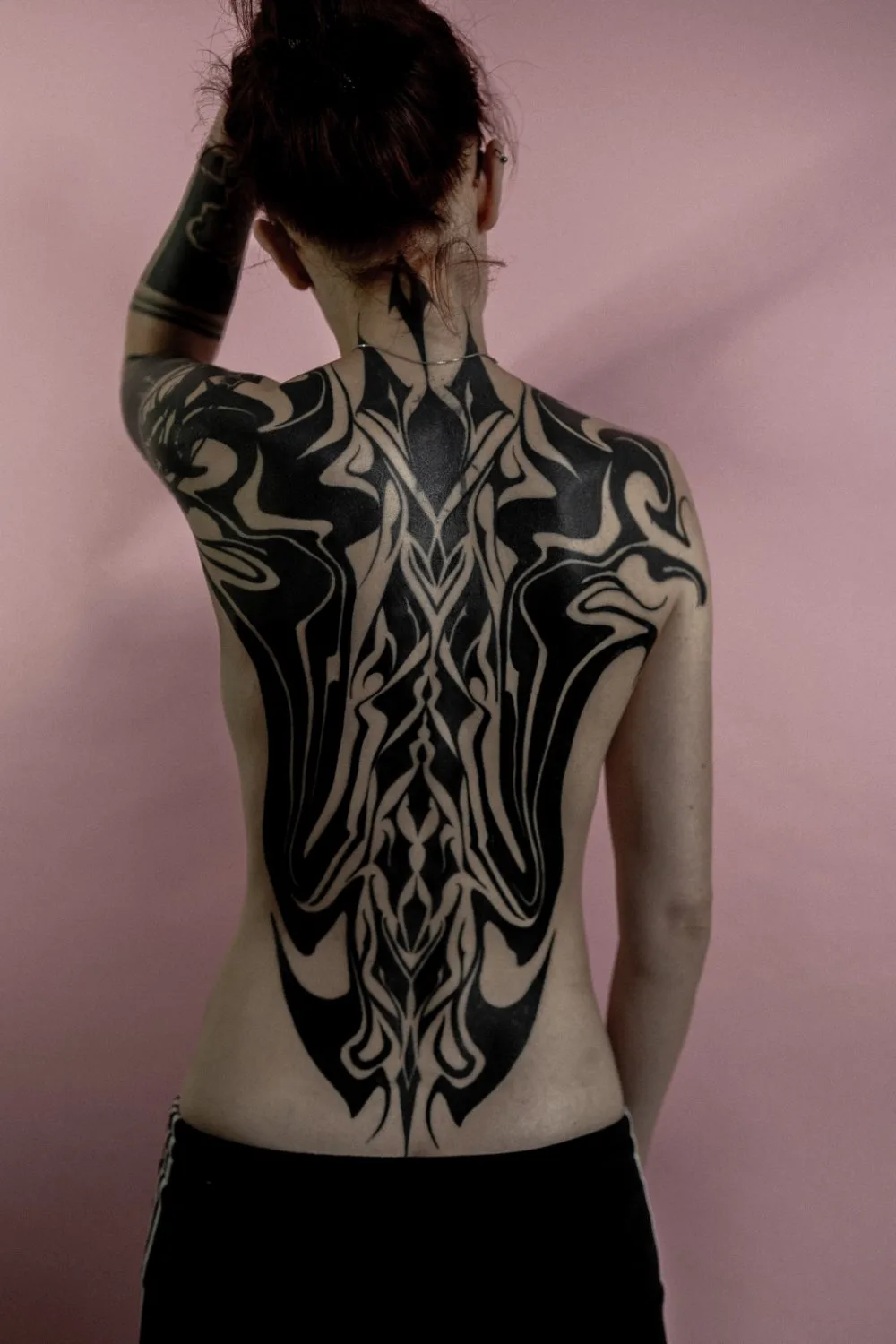 A person viewed from behind with extensive black abstract tattoos covering the back, shoulders, and upper arms. The design is symmetrical and bold, with sharp angular points and sweeping curves in solid black ink against fair skin and a pink background.
