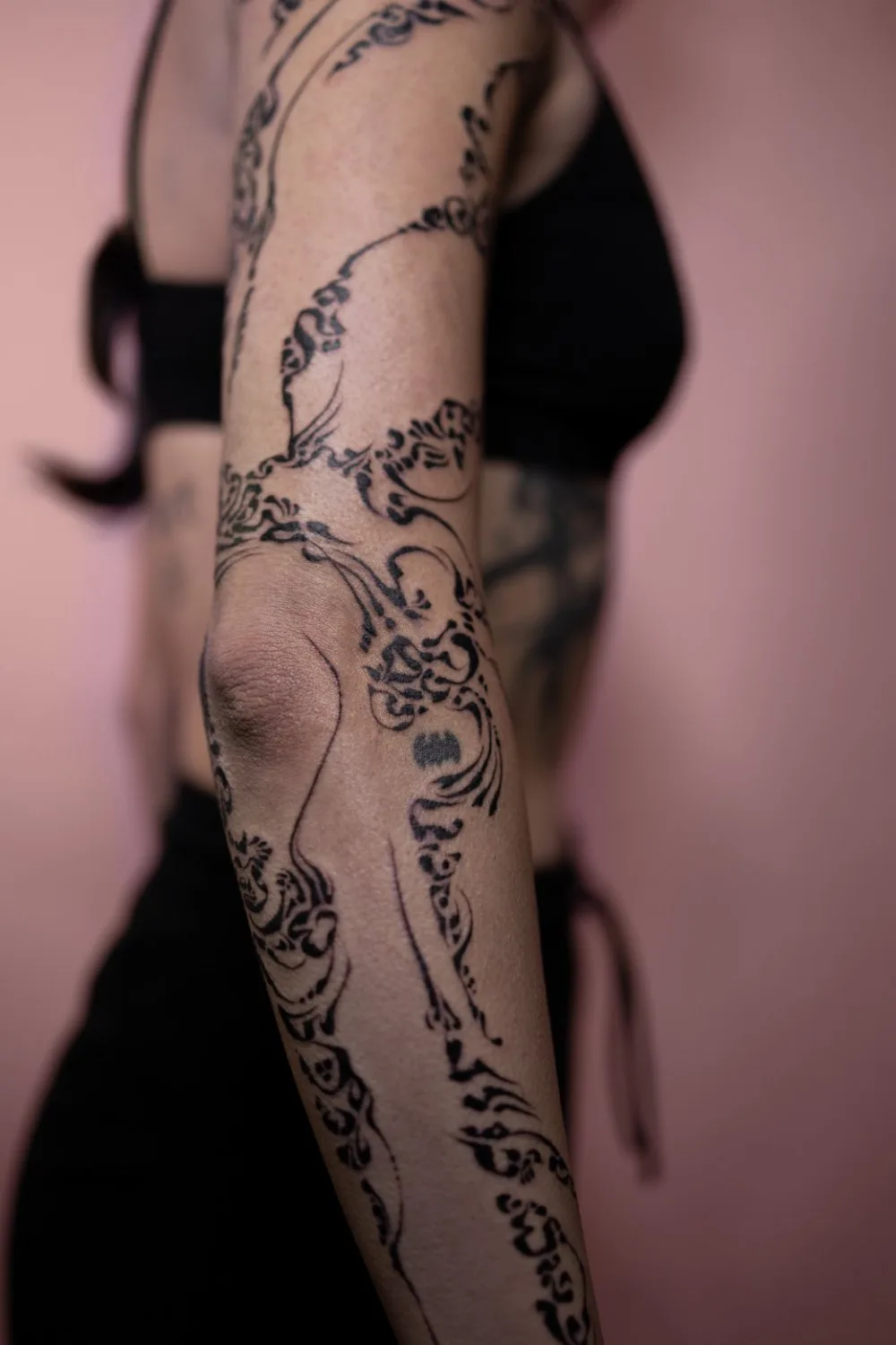 A close-up profile view of a person's upper arm showing a flowing black ink tattoo that extends from the shoulder to the elbow. The design features thin, precise lines with some thicker filled areas, wrapping organically around the arm. The background is soft pink.