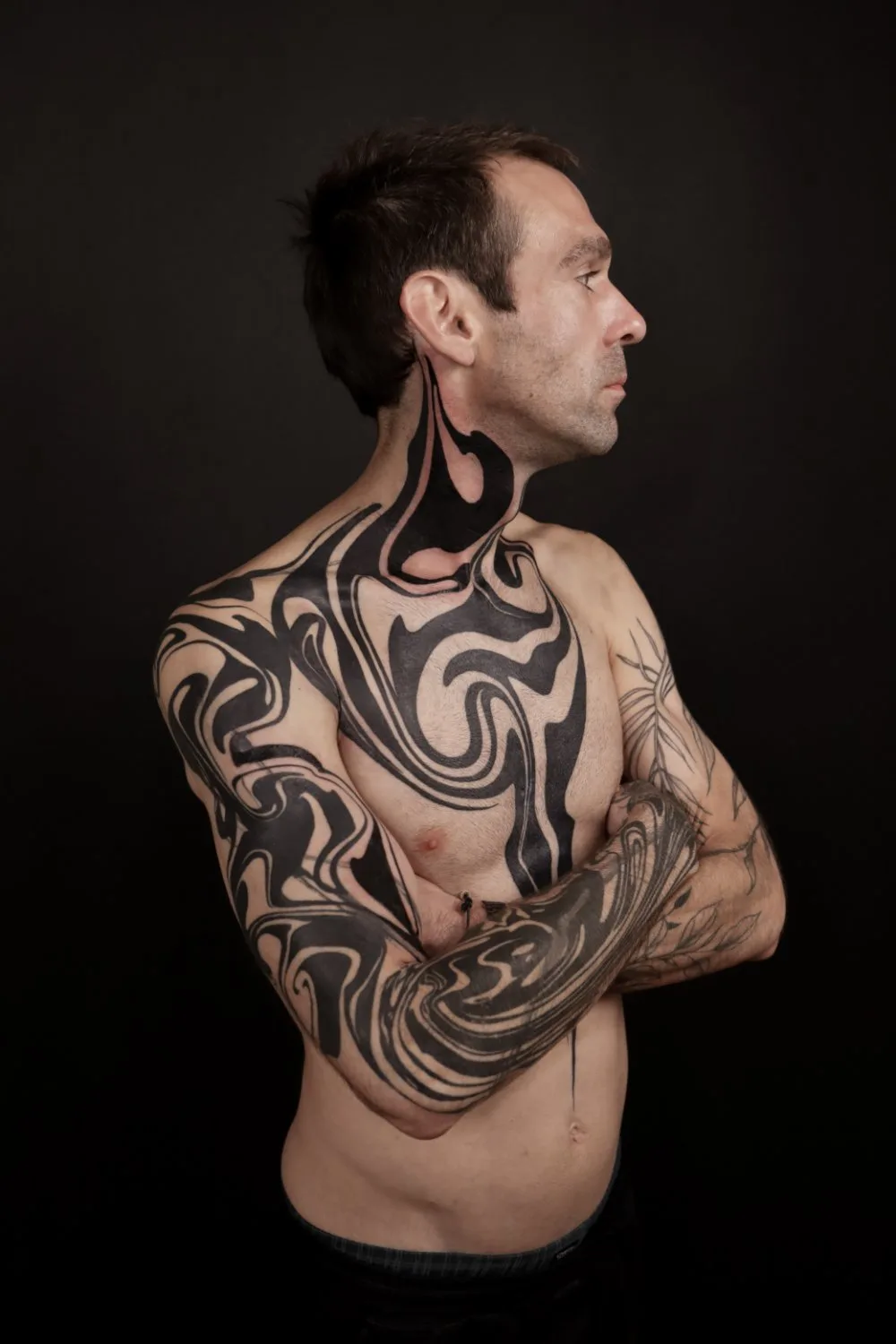 A bare-chested man in profile with extensive black ink tattoos covering his neck, chest, and arms. The designs are abstract and organic, with swirling lines of varying thickness. His arms are crossed, and the background is dark.