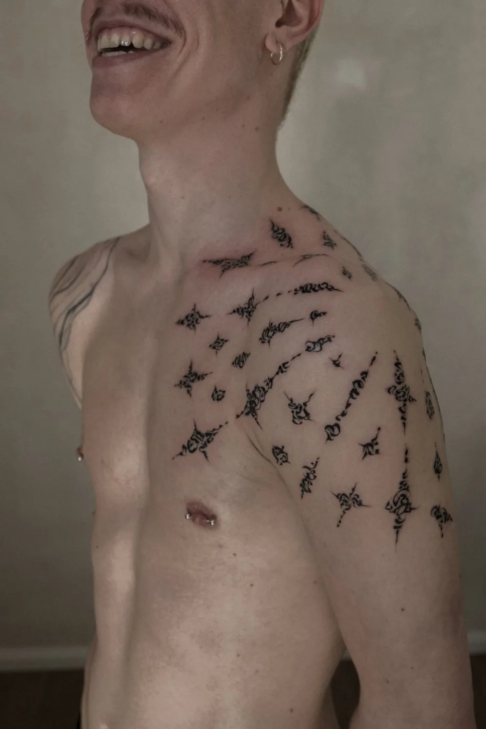 A person's upper torso and shoulder showing intricate black ink tattoos covering the left shoulder, collarbone, and chest. The design features fine linework and solid black fills in abstract ornamental patterns. The person is smiling and looking upward.