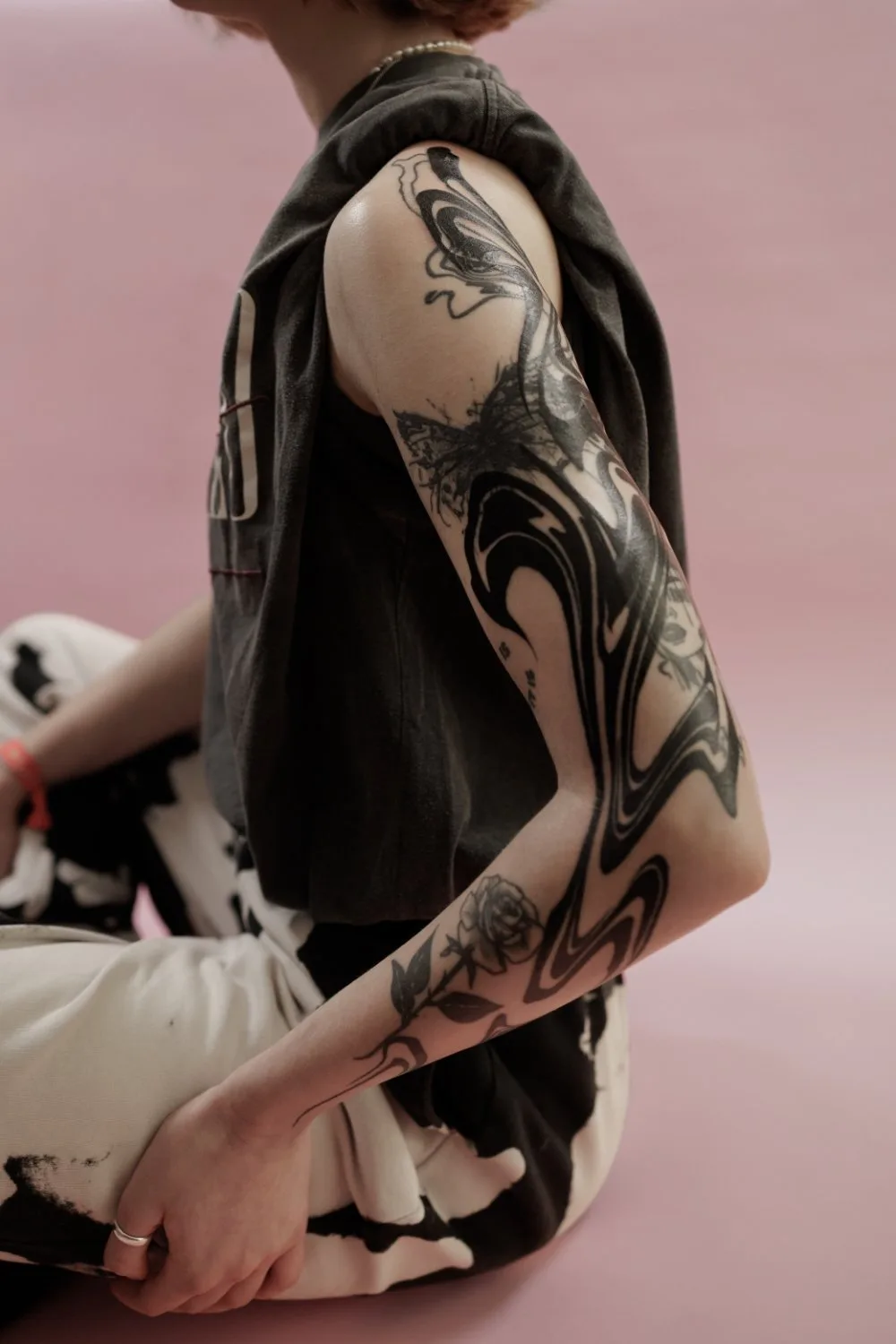 A person's right arm and shoulder showing elaborate black ink tattoos. The upper arm features large, fluid abstract shapes in solid black, while the lower forearm shows a rose tattoo in black and grey. The person wears a grey hooded garment draped over the shoulder. Background is soft pink.