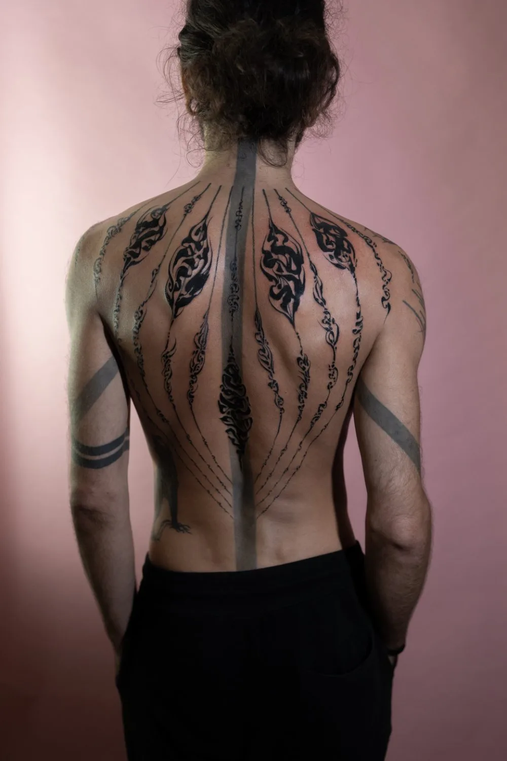 A person viewed from behind with extensive black ink tattoos covering the back, shoulders, and upper arms. The design radiates outward from a central vertical line along the spine, with organic curving motifs and varying line weights. The person wears black pants and the background is soft pink.