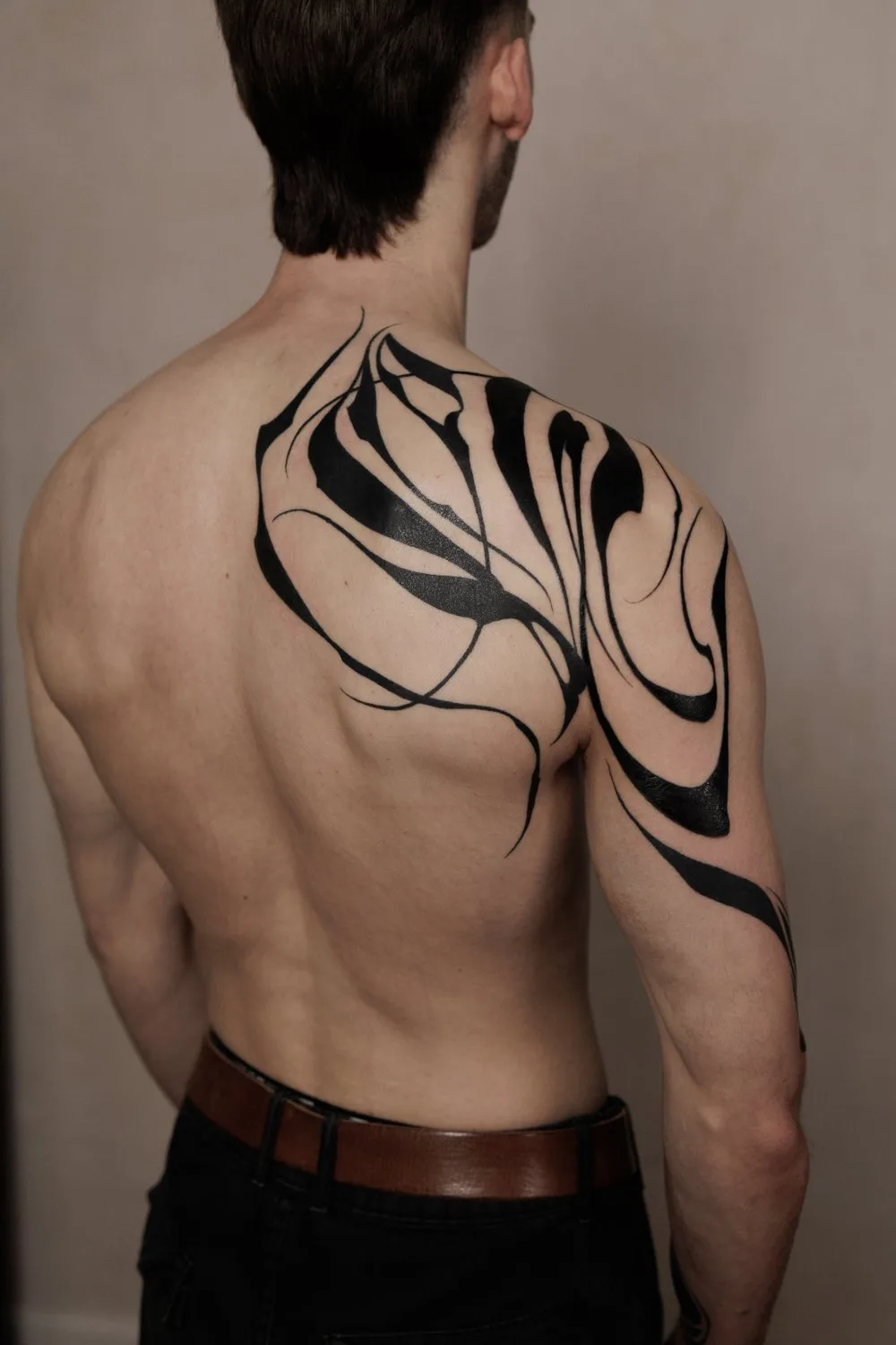 The upper back and left shoulder of a shirtless man showing a large abstract black tattoo. The design combines thick, solid black shapes with thinner swirling lines, creating a dynamic organic composition that extends from the shoulder blade across the upper arm.