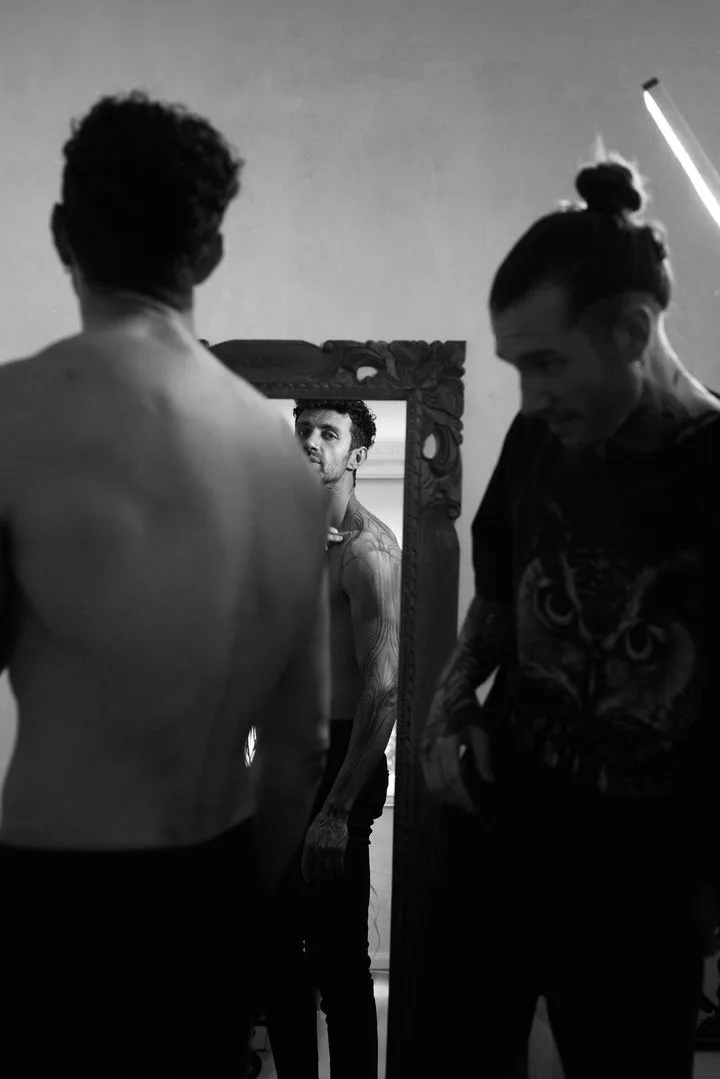 Black and white photograph of three men in an indoor setting. One man faces a large ornate mirror with his back to the camera; in the mirror's reflection a second heavily tattooed bare-chested man looks directly at the viewer. A third man, wearing a t-shirt with an owl graphic, stands partially out 