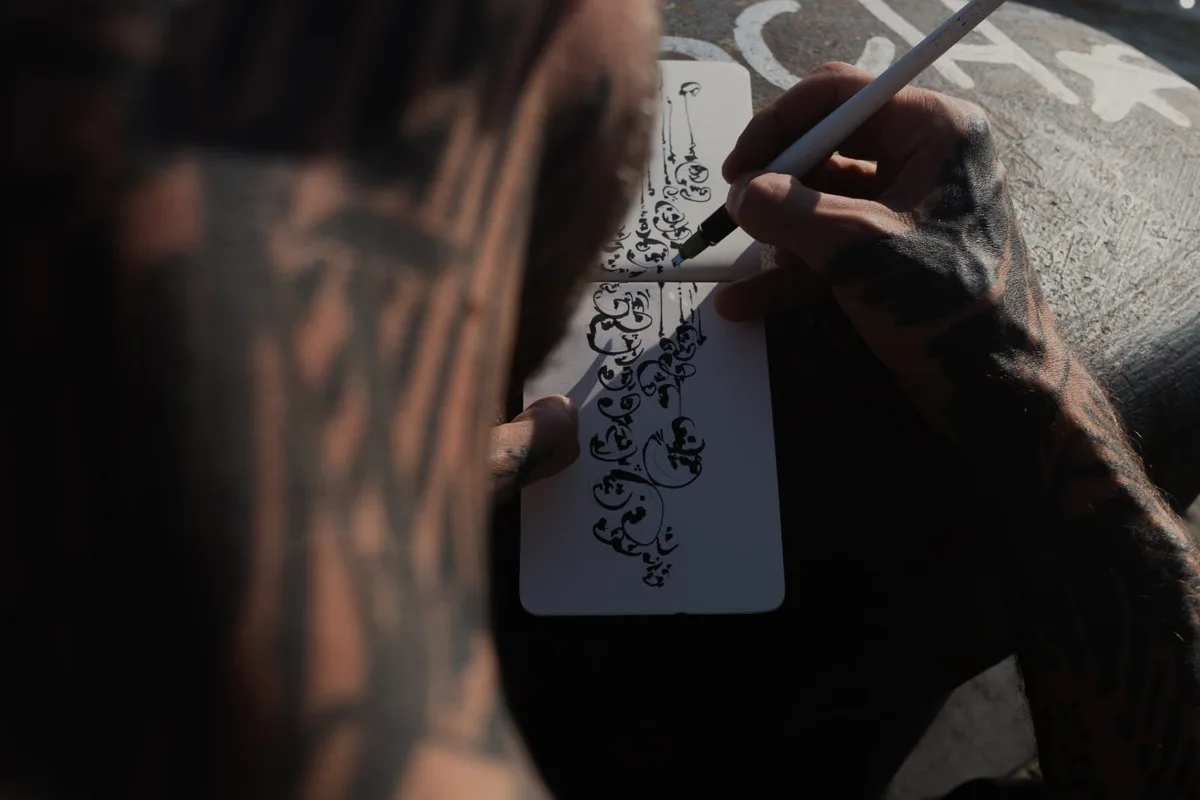 A high-angle close-up of tattooed hands drawing intricate abstract black ink designs in a small white notebook. The drawing features flowing loops and calligraphic marks. The person's heavily tattooed arms are visible, and the background is a dark textured surface.