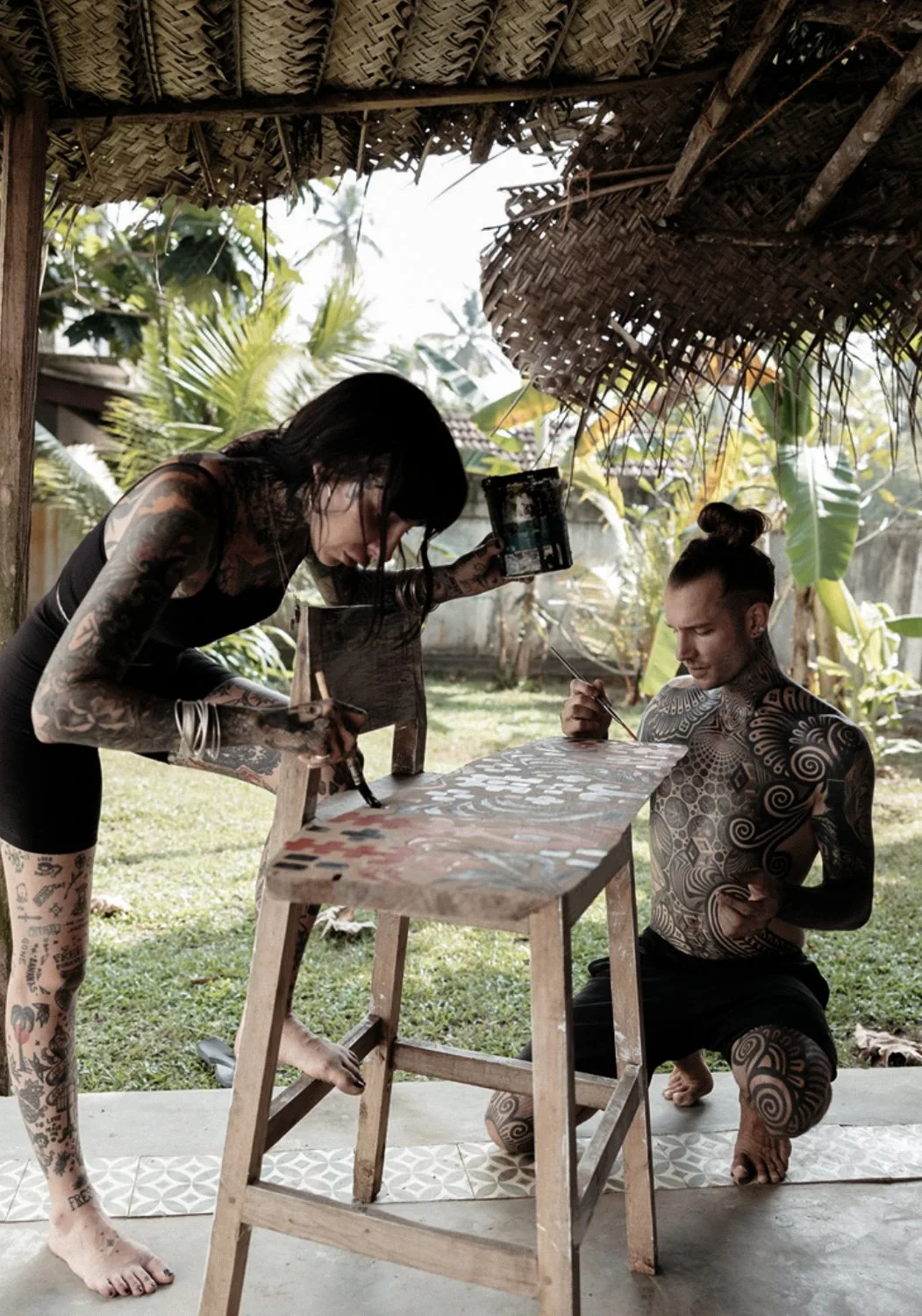A woman and a shirtless man, both heavily tattooed, painting a wooden stool in an outdoor tropical setting with a thatched roof. The woman holds a paint can and brush; the man kneels beside the stool. Lush greenery is visible in the background.